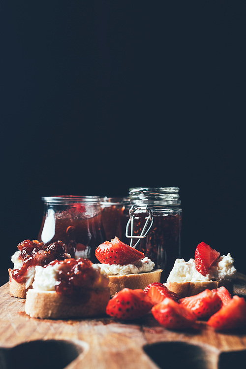 selective focus of sandwiches with cream cheese, strawberry slices and fruit jam on cutting board on black