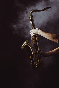 cropped shot of musician holding saxophone in smoke on black