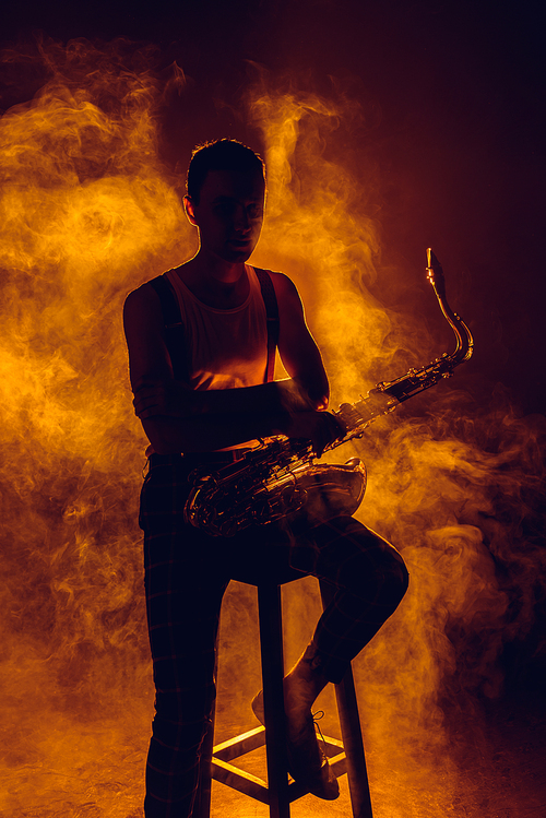 silhouette of young jazzman sitting on stool and holding saxophone in smoke