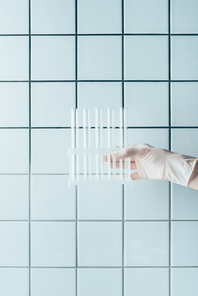 cropped shot of doctor in glove holding test tubes in stand in front of tiled white wall