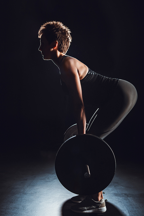 side view of female athlete doing exercise with barbell at gym, black background
