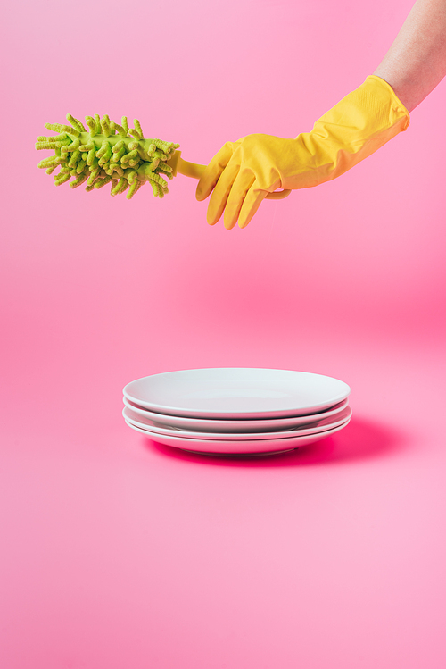 cropped image of woman in rubber glove holding dish microfiber brush over stack of white plates, pink background