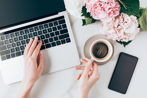 cropped view of woman using laptop and holding cup of coffee on tabletop with smartphone and hortensia flowers