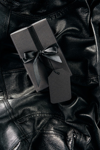 top view of black wrapped gift with blank price tag on black leather jacket background, black friday concept