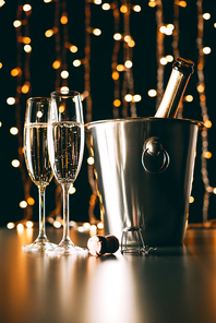 champagne bottle in bucket and glasses on garland light background, christmas concept