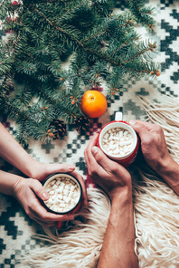cropped shot of couple holding cups of cocoa hot drinks at background with pine tree branches and tangerine