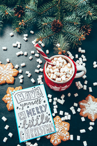 flat lay with cup of hot drink with marshmallow, cookies and postcard with have yourself merry little christmas lettering on black tabletop