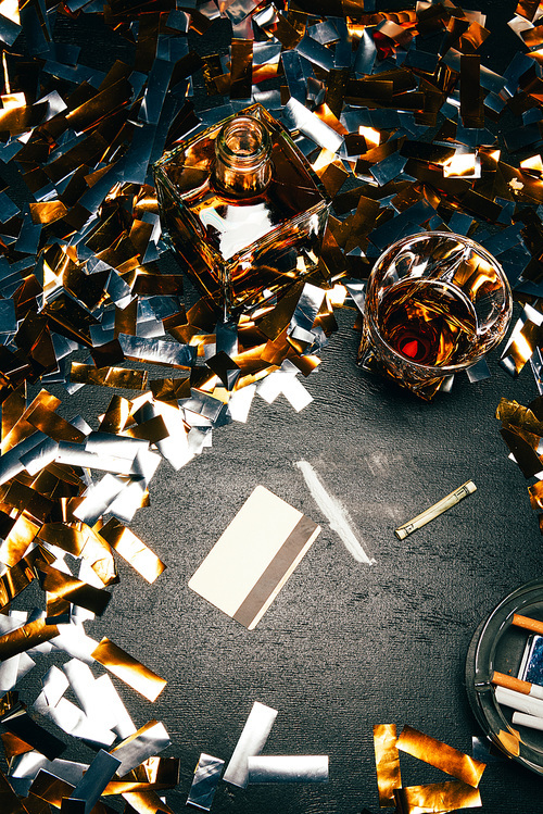 top view of whiskey, rolled banknote, credit card and cocaine on table covered by golden confetti