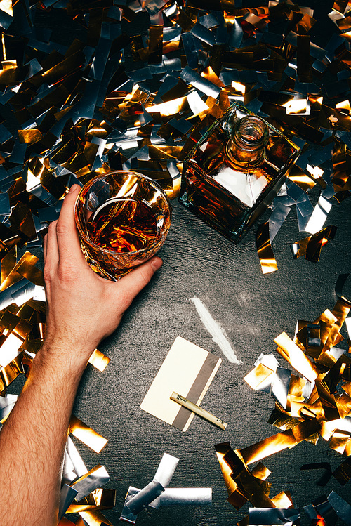 partial view of man with whiskey glass near cocaine with rolled banknote and credit card on table covered by golden confetti