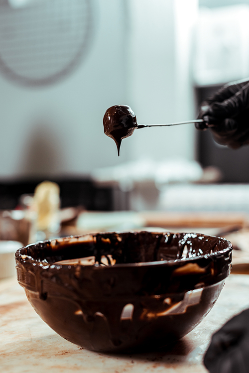 cropped view of chocolatier in black latex glove holding stick with tasty candy near chocolate in bowl