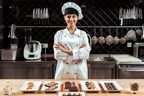 happy chocolatier in chef hat standing with crossed arms near tasty chocolate candies on plates