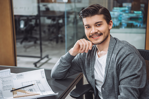 handsome young architect sitting at workplace with building plans at office
