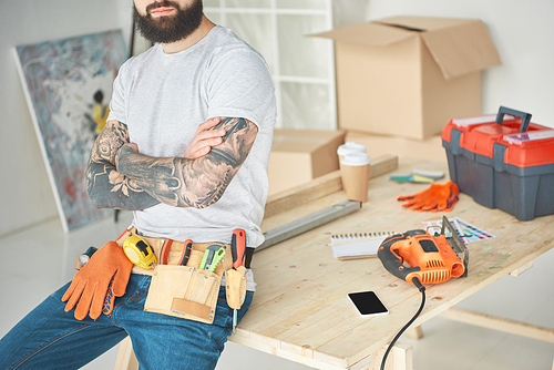 cropped shot of bearded tattooed man with tool belt sitting with crossed arms on wooden table