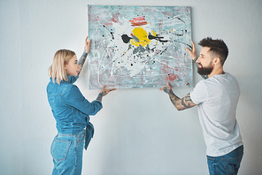 young tattooed couple hanging painting on wall together at new apartment