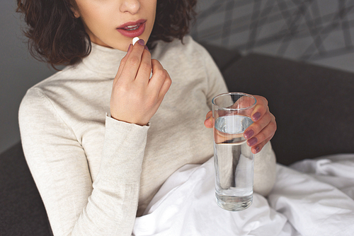 cropped image of sick woman taking pill and holding glass of water at home
