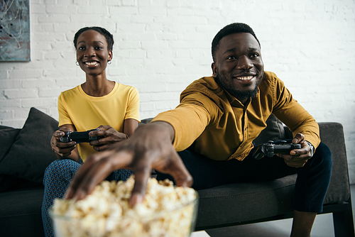 happy young african american couple eating popcorn and playing with joysticks at home