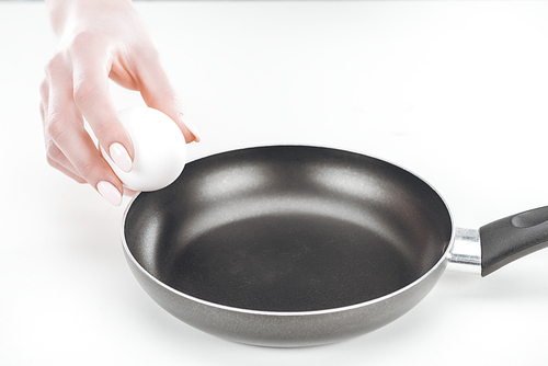 partial view of woman holding chicken egg near pan on white background