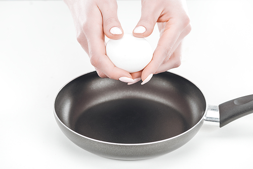 cropped view of woman holding chicken egg in hands near pan on white background