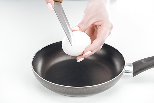 cropped view of woman holding chicken egg and knife near pan on white background
