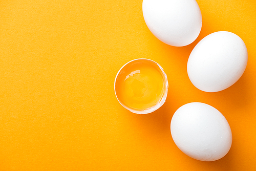 top view of smashed chicken egg with yolk on bright orange background among white whole eggs