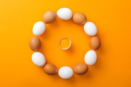top view of white and brown organic chicken eggs arranged in round frame with smashed one inside on bright orange background