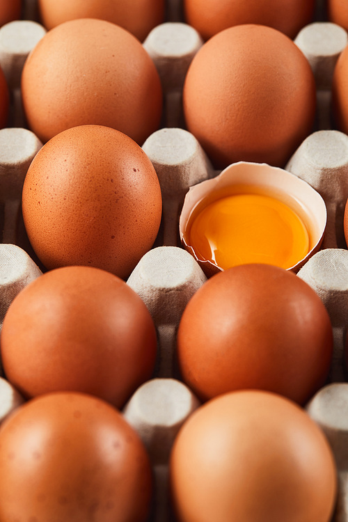 selective focus of broken eggshell with yellow yolk near eggs in carton box