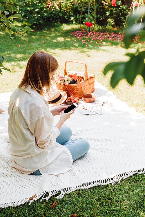 blonde girl sitting on blanket in garden and having picnic at sunny day while using smartphone