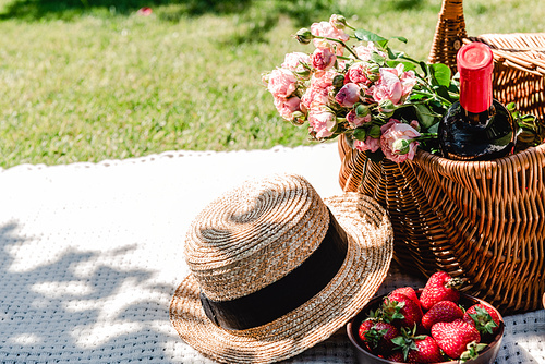 wicker basket with roses and bottle of wine on white blanket near straw hat and strawberries at sunny day in garden