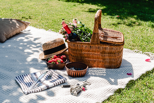 wicker basket with roses and wine on white blanket near straw hat, cutlery on napkin and berries at sunny day in garden