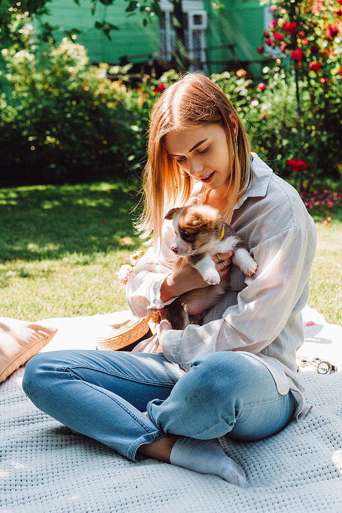 blonde girl sitting on blanket in garden with crossed legs and adorable puppy at sunny day