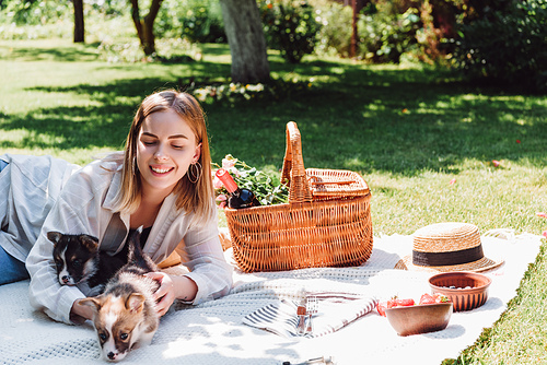 smiling blonde girl sitting on blanket in garden and having picnic with puppies at sunny day