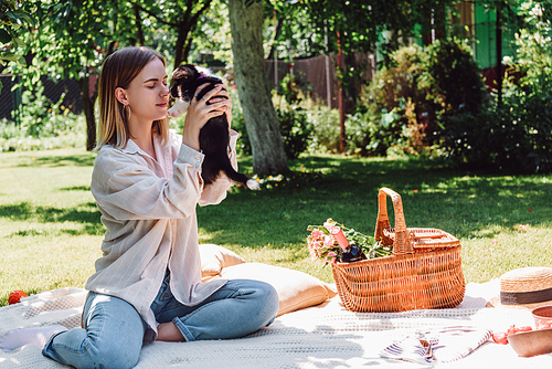 blonde girl sitting on blanket in garden and holding puppy at sunny day