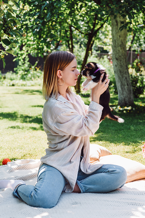 blonde girl with closed eyes holding cute puppy while sitting in green garden