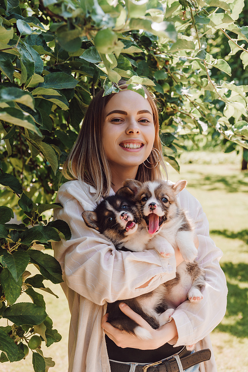 happy blonde girl holding cute puppies in garden near green tree
