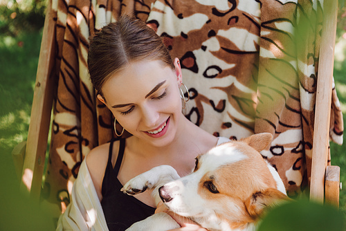 selective focus of green leaves and happy blonde girl holding corgi dog while sitting in deck chair in garden