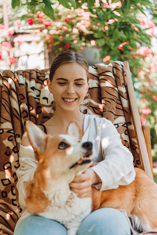 selective focus of happy blonde girl holding corgi dog while sitting in deck chair in garden