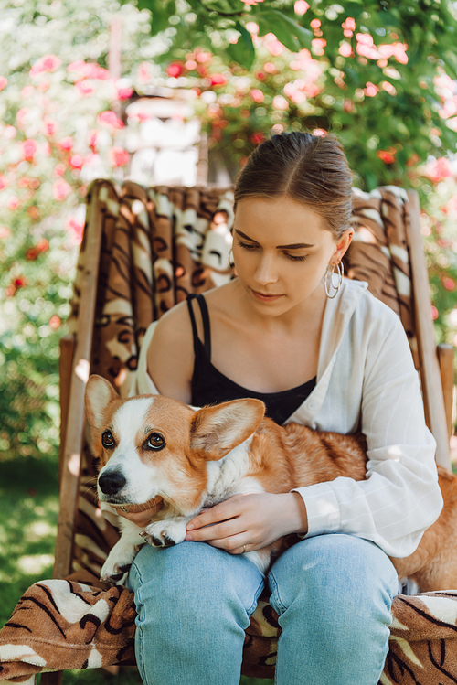 blonde girl holding corgi dog while sitting in deck chair in green garden