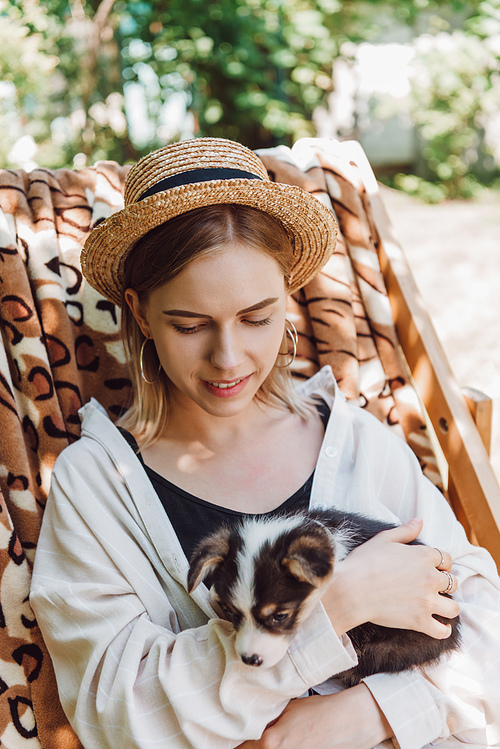 smiling blonde girl in straw hat holding puppy while sitting in deck chair in garden