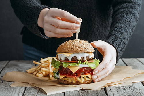 cropped view of female hands with big tasty cheeseburger and french fries on baking paper