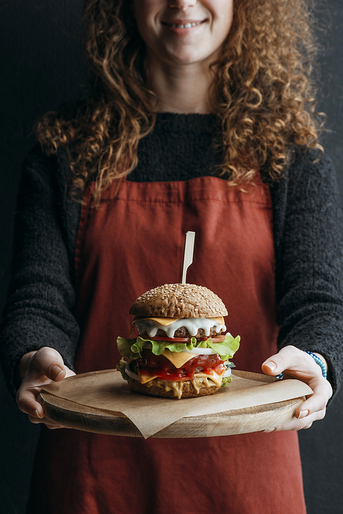 cropped view of girl in apron holding wooden board with big homemade cheeseburger