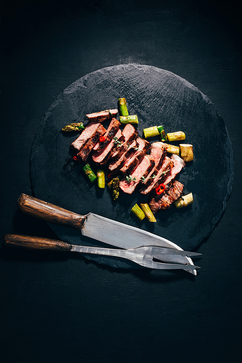 top view of delicious sliced grilled meat with asparagus| meat fork and knife on black slate board