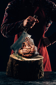 cropped shot of chef in apron holding meat knife and delicious grilled ribs on wooden stump