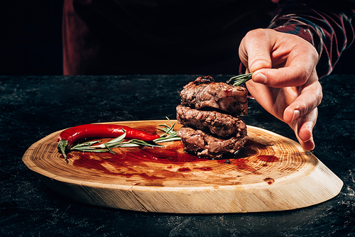 cropped shot of person putting rosemary on grilled steaks with chili pepper and sauce on wooden board