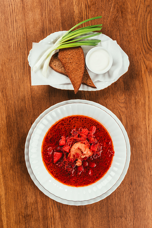 Top view of borscht dish in white plate served with rye bread on wooden table