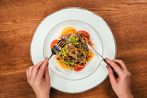 Top view of woman eating fresh vegetable salad served on white plate on wooden table