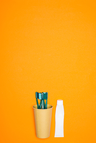 top view of toothbrushes in paper cup and tube of toothpaste| isolated on orange