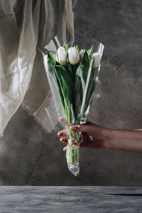cropped view of woman holding bouquet of tulips in cellophane on grey surface
