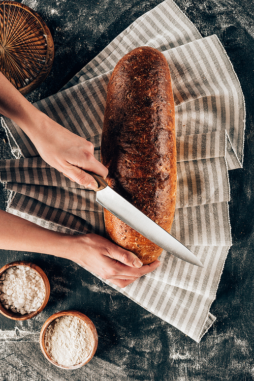 cropped shot of woman cutting loaf of bread on linen on dark tabletop with flour