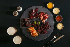 top view of gourmet assorted meat on slate board| various sauces and glasses of beer on black