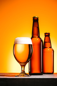 close up view of bottles and glass of beer with foam on surface on orange background